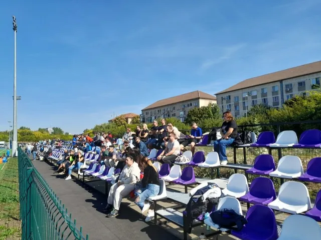Oamenii stau așezați pe scaune de culoare albastră și albă în tribunele unui stadion sub un cer senin.