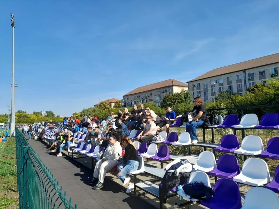 Oamenii stau așezați pe scaune de culoare albastră și albă în tribunele unui stadion sub un cer senin.