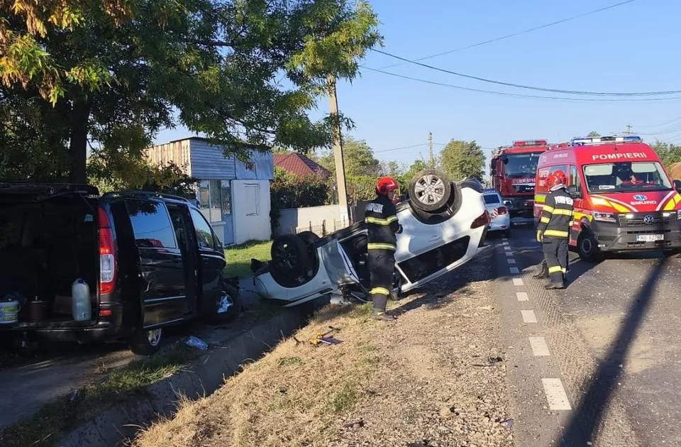 Un vehicul alb este răsturnat pe o parte pe marginea drumului, lângă un vehicul negru și două autospeciale de pompieri.