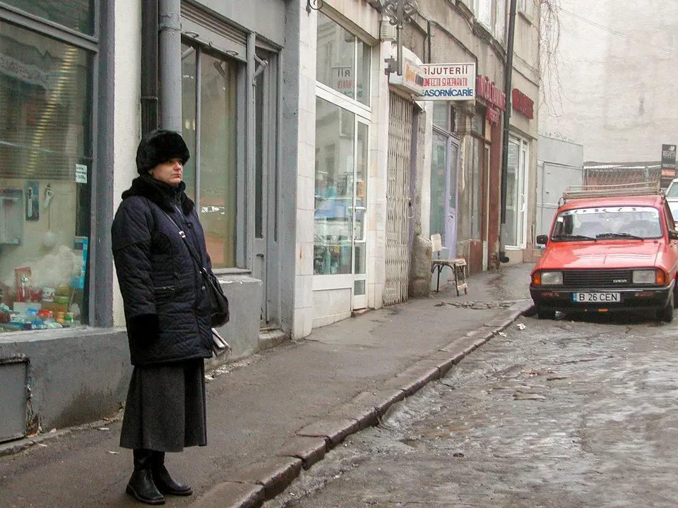 O femeie cu o haină neagră și o fustă gri stă pe un trotuar lângă vitrinele unor magazine de pe o stradă îngustă.