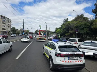 O mașină de poliție locală albă cu marcaje și lumină de semnalizare este oprită pe un drum asfaltat lângă alte vehicule și un autobuz.