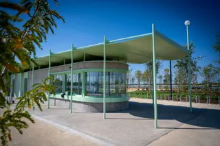O clădire circulară cu ferestre mari de sticlă este acoperită de un copertină verde deschisă susținută de stâlpi sub un cer senin.