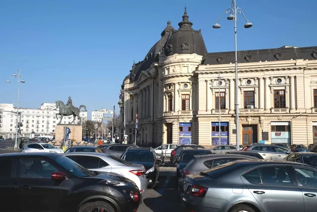 Mașini sunt parate pe un drum în fața unei clădiri mari, de stil clasic, sub un cer senin.