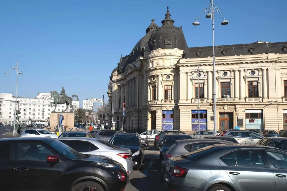 Mașini sunt parate pe un drum în fața unei clădiri mari, de stil clasic, sub un cer senin.