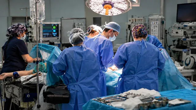 O echipă de medici în halate și căciuli sterile execută o intervenție chirurgicală într-o sală de operații dotată cu echipamente medicale și lumini puternice.