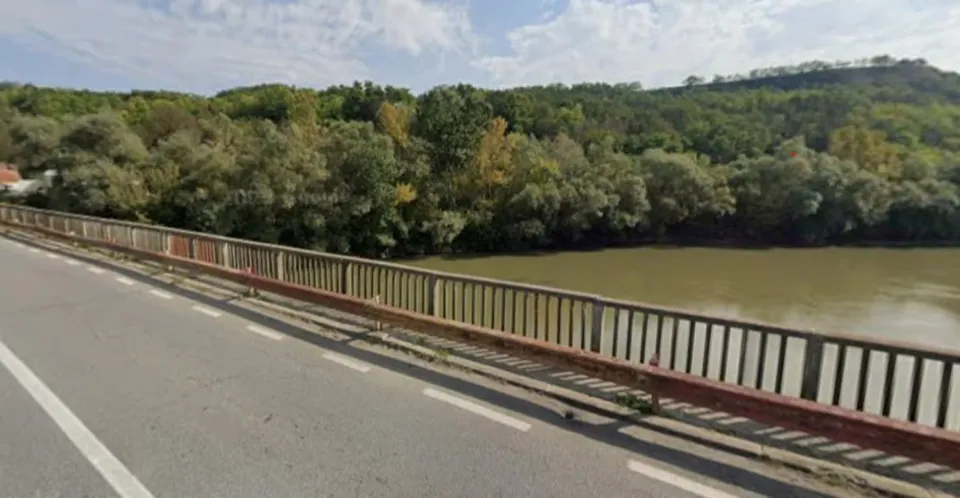 O porțiune de drum asfaltat cu linii albe este delimitată de un parapet metalic deasupra unui râu cu apă de culoare maronie sub un cer înnorat.