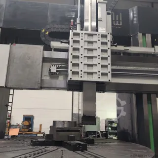 O mașină de lucru industrială mare cu o componentă metalică dreptunghiulară suspendată deasupra unei platforme rotative într-un atelier.