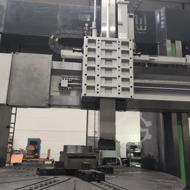 O mașină de lucru industrială mare cu o componentă metalică dreptunghiulară suspendată deasupra unei platforme rotative într-un atelier.
