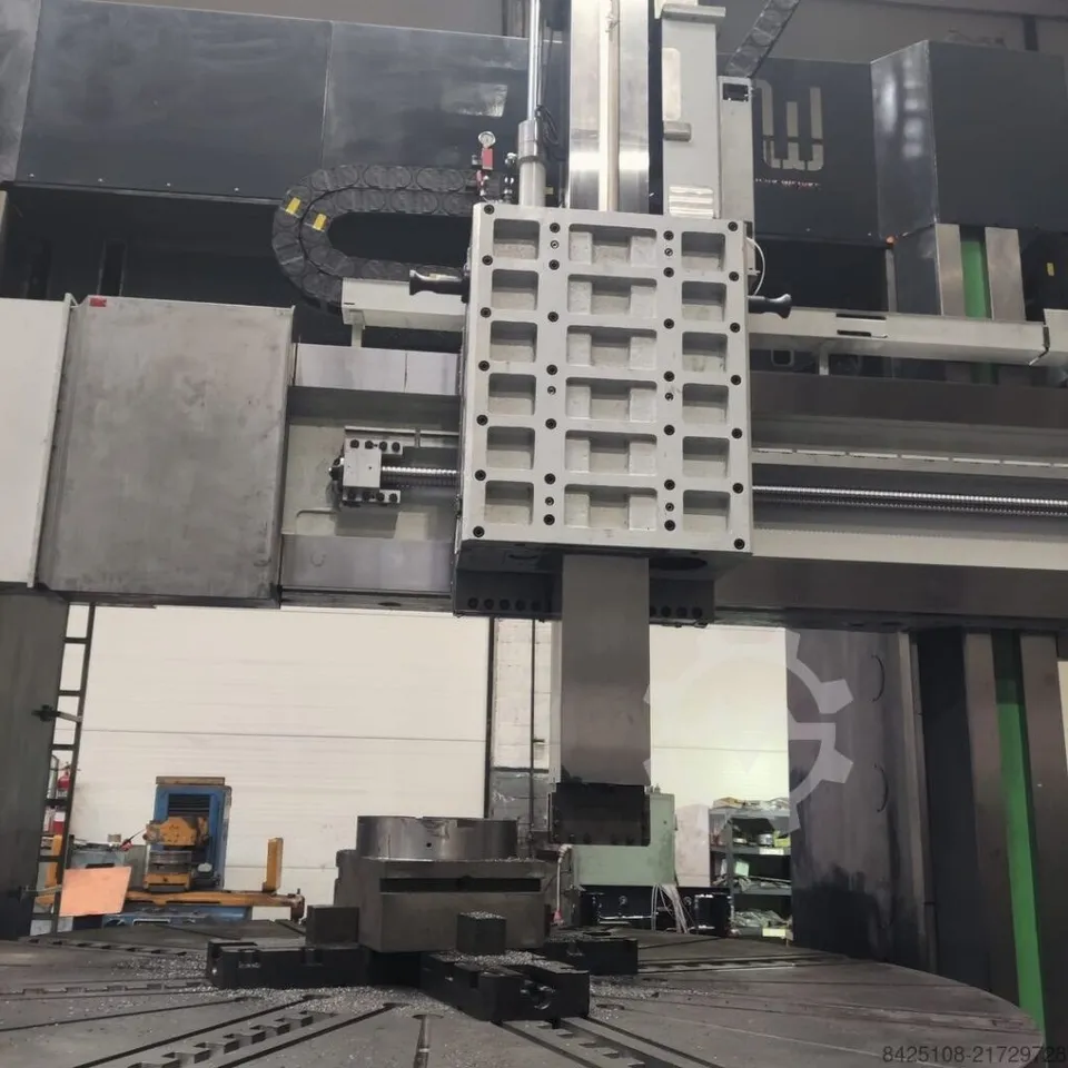 O mașină de lucru industrială mare cu o componentă metalică dreptunghiulară suspendată deasupra unei platforme rotative într-un atelier.