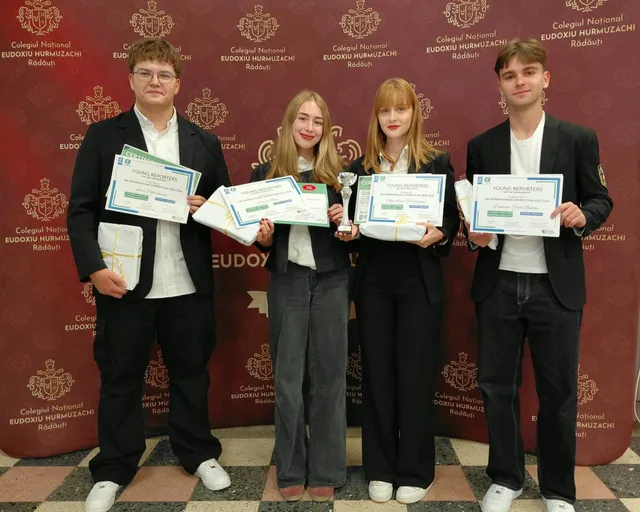 Patru persoane stau în picioare în fața unui fundal roșu, ținând diplome și un trofeu argintiu.