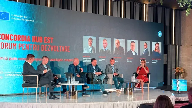 Șase persoane stau așezate pe scaune pe o scenă în fața unui ecran mare care afișează portrete și text colorat.
