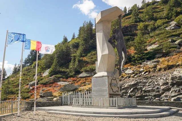 Un monument sculptat din piatră deschide o figură umană sub un element geometric înalt, situat pe un soclu lângă trei steaguri.