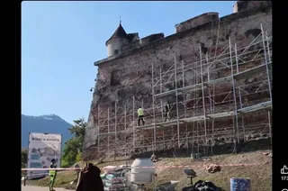 O clădire veche din piatră este acoperită parțial de niște schele metalice, în timp ce un om în vestă reflectorizantă se află pe acestea.