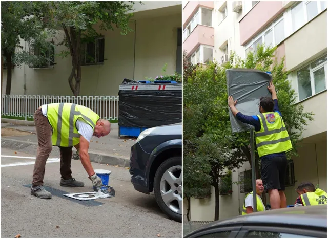Un bărbat cu vestă reflectorizantă galbenă este aplecat lângă un marcaj pe asfalt, în timp ce un alt bărbat instalează un panou gri pe un stâlp.