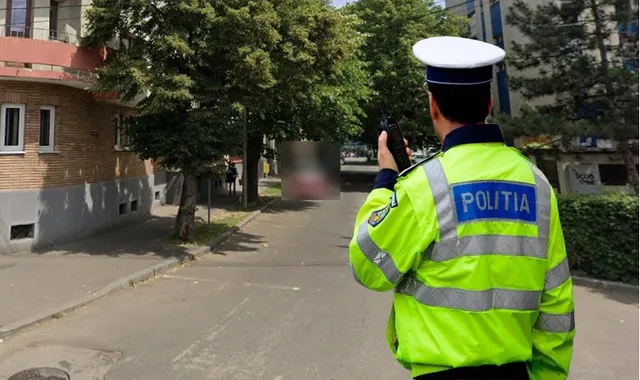 Un politist în uniformă luminoasă portocalie stă cu spatele, privind spre un drum cu copaci și clădiri.