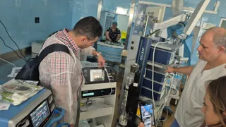 Un bărbat într-un halat de unică folosință și un alt bărbat în tricou alb privesc un aparat medical într-o încăpere cu pereți albaștri.