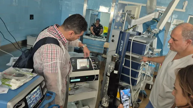 Un bărbat într-un halat de unică folosință și un alt bărbat în tricou alb privesc un aparat medical într-o încăpere cu pereți albaștri.