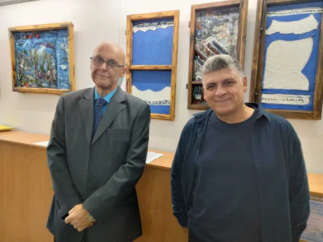 Un bărbat cu ochelari și sacou gri stă lângă un bărbat cu tricou negru și sacou albastru în fața unor tablouri înrame în lemn.