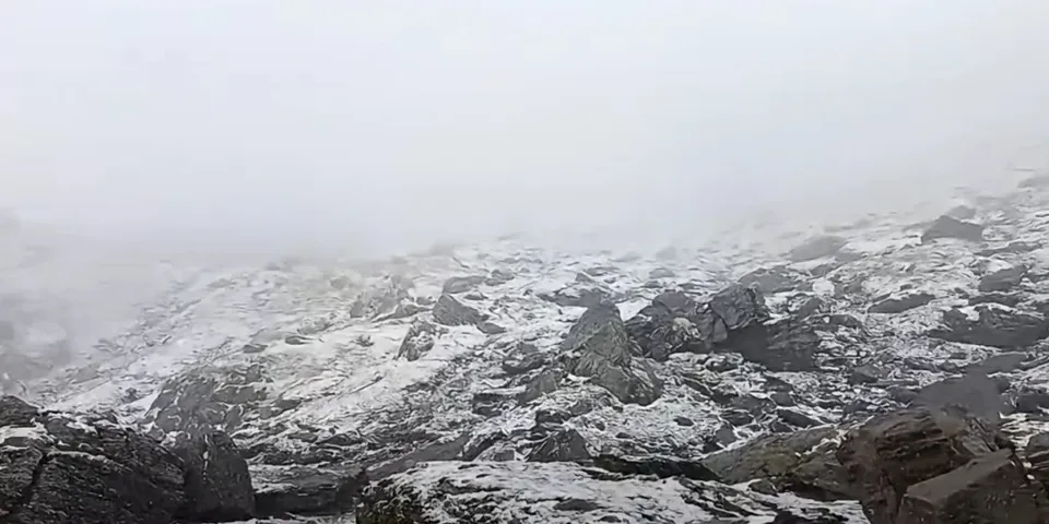 Un peisaj muntos acoperit de zăpadă și pietre întinse sub un cer senin și brumat.