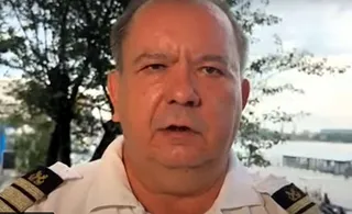 Un bărbat cu uniformă albă și epoleți negri cu dungi aurii privește spre cameră în fața unor copaci și a unui cer deschis.