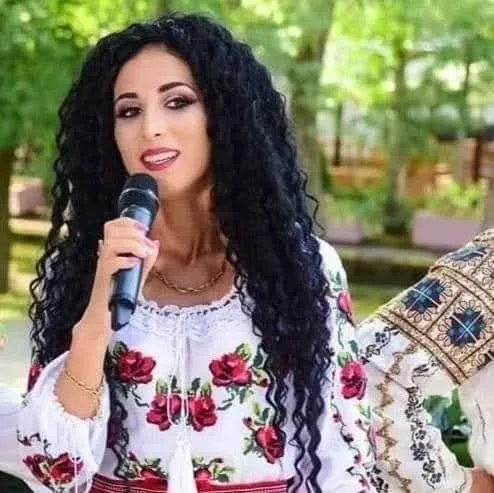 O femeie cu părul negru și creț îl ține pe o femeie cu o bluză tradițională albă brodată cu flori roșii și un microfon negru.