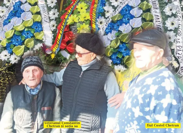 Trei persoane vârstnice stau în fața unor corbuțe de flori colorate, purtând haine de iarnă și pălării.