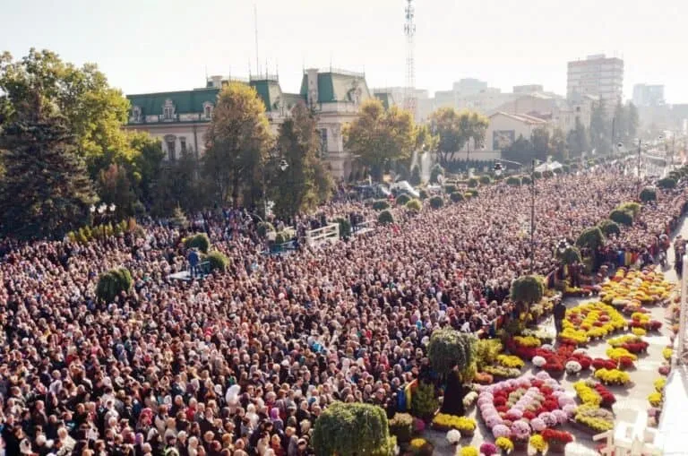 O mulțime mare de oameni este adunată pe o stradă lată lângă straturi de flori colorate și copaci decorați.