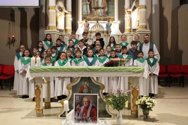 Un grup mare de copii îmbrăcați în robe albe cu panglici verzi stau în grup în fața unui altar într-o biserică.