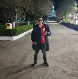 Un bărbat cu ochelari de soare, purtând un tricou roșu și negru, o jachetă neagră și pantaloni închiși, stă pe un drum pietruit noaptea.