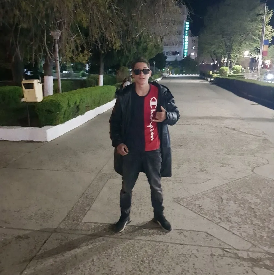 Un bărbat cu ochelari de soare, purtând un tricou roșu și negru, o jachetă neagră și pantaloni închiși, stă pe un drum pietruit noaptea.