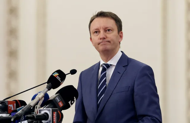 Un bărbat într-un costum albastru și cravată în dungi stă în fața mai multor microfoane de presă.