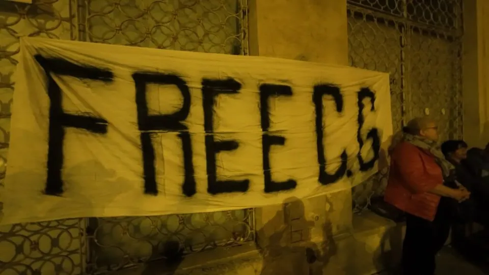 Un banner alb cu textul negru scris de mână este întins pe un gard metalic în fața unei clădiri noaptea.