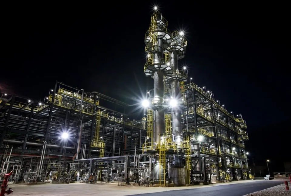 O platformă industrială iluminată noaptea prezintă turnuri înalte, structuri metalice și conducte complexe sub un cer negru.