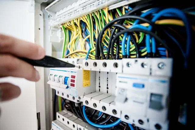 Mâini ținând un cablu negru în apropierea unui panou electric cu diverse fire colorate și întrerupătoare.