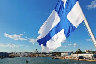O steag finlandez albastru și alb flutură pe un stâlp sub un cer senin deasupra unui port cu clădiri și bărci.