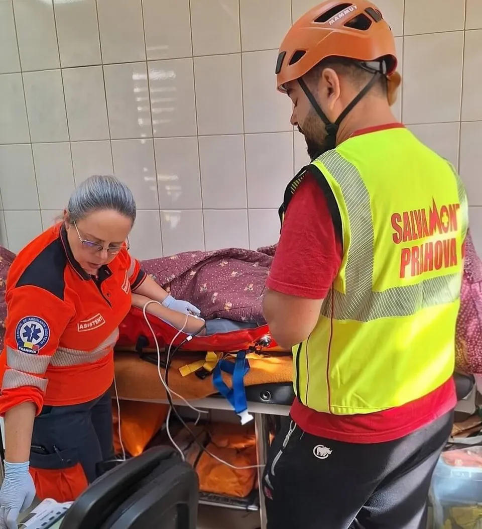Un bărbat cu cască portocalie și vestă reflectorizantă privește o femeie în echipament portocaliu care manipulează un obiect pe un pat medical.