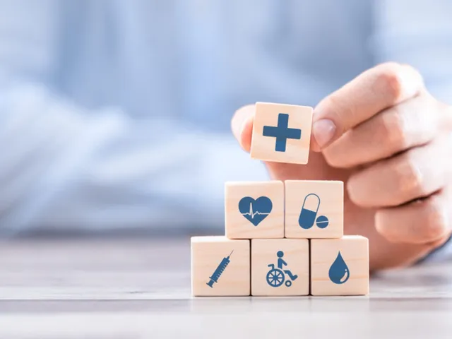 O mână ține un cub de lemn cu o cruce albastră peste o piramidă de cuburi de lemn cu diverse pictograme medicale pe fundal albastru.