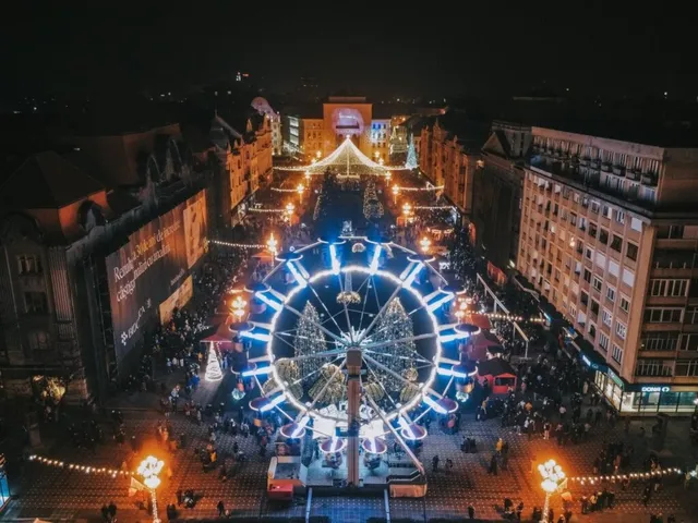 O roată romantică iluminată cu lumini albastre și albe strălucitoare se află într-o piață urbană plină de oameni și standuri de vânzare noaptea.