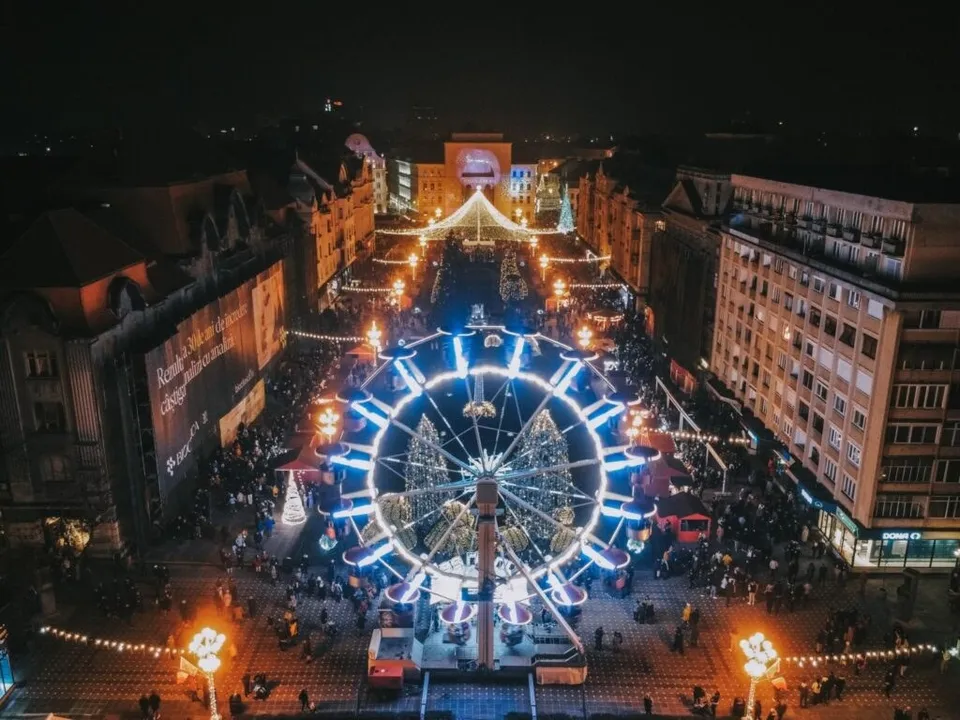 O roată romantică iluminată cu lumini albastre și albe strălucitoare se află într-o piață urbană plină de oameni și standuri de vânzare noaptea.