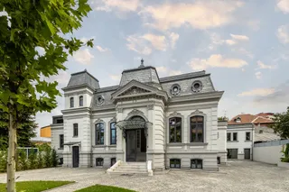 O clădire albă cu acoperișuri înclinate și o curte pavată, cu arbori în stânga și cer parțial noros.