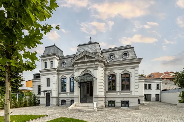 O clădire albă cu acoperișuri înclinate și o curte pavată, cu arbori în stânga și cer parțial noros.
