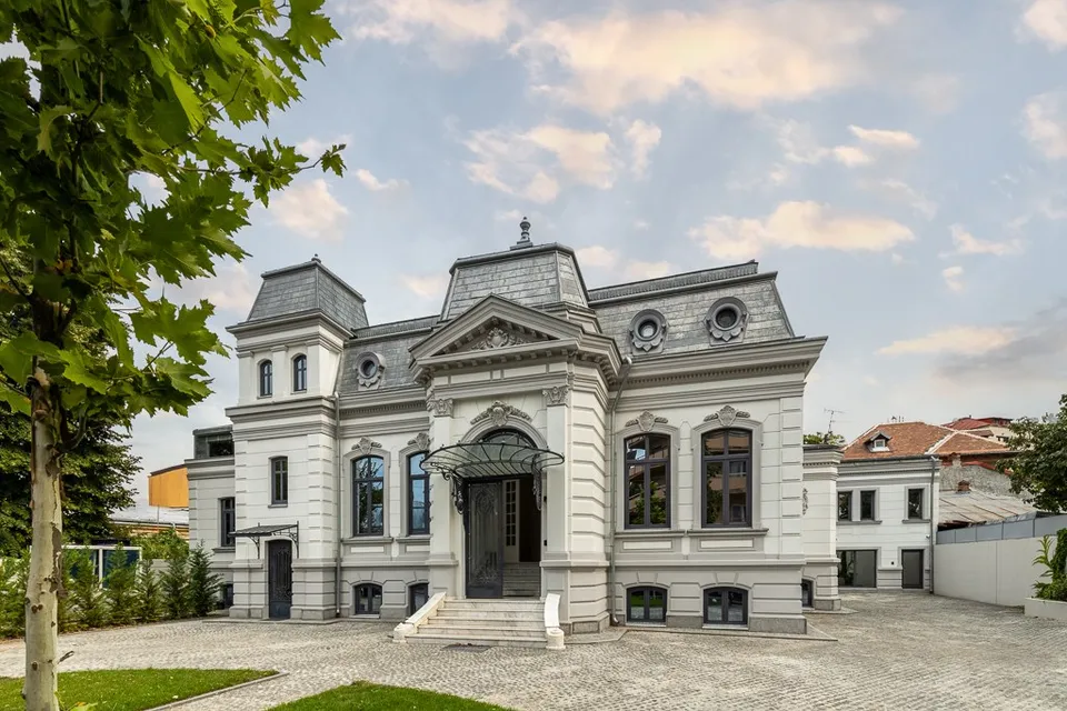 O clădire albă cu acoperișuri înclinate și o curte pavată, cu arbori în stânga și cer parțial noros.