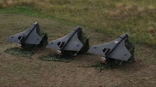 Trei drone gri sunt așezate pe un câmp de iarbă, acoperite parțial de plasă de camuflaj verde.