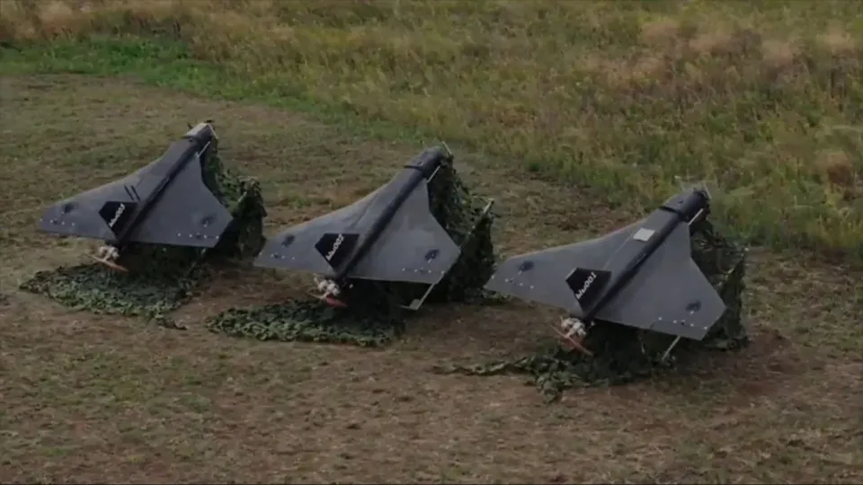 Trei drone gri sunt așezate pe un câmp de iarbă, acoperite parțial de plasă de camuflaj verde.