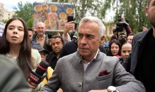 Un bărbat cu părul cărunt și sacou gri stă în mijlocul unei mulțimi de oameni, fiind înconjurat de microfoane și camere de filmat.
