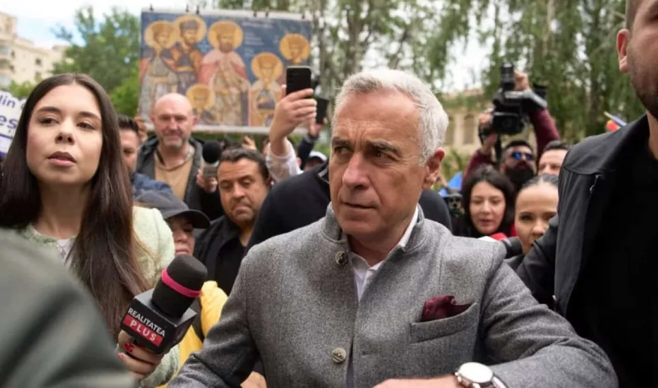 Un bărbat cu părul cărunt și sacou gri stă în mijlocul unei mulțimi de oameni, fiind înconjurat de microfoane și camere de filmat.