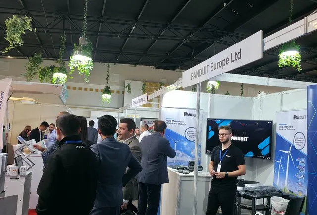 Un grup de oameni stă în picioare în fața unui stand de expoziție cu panouri informative și un ecran digital.