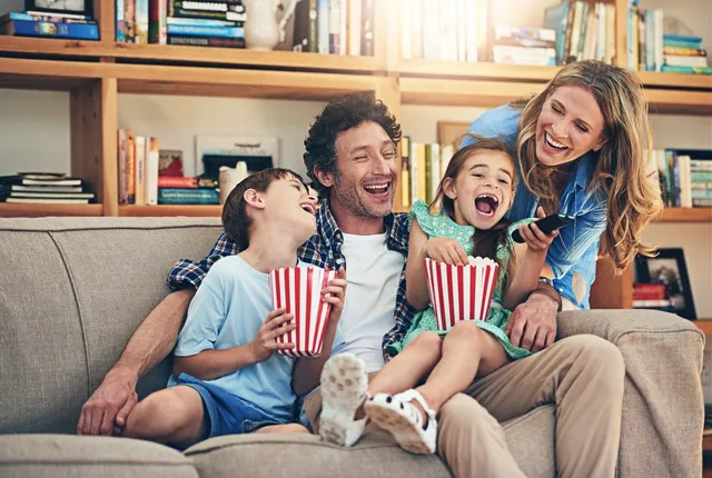 O familie de patru persoane zâmbește și râde pe o canapea, având în mână boluri cu popcorn.