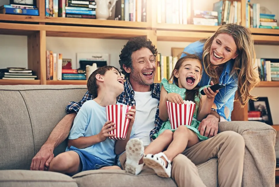 O familie de patru persoane zâmbește și râde pe o canapea, având în mână boluri cu popcorn.