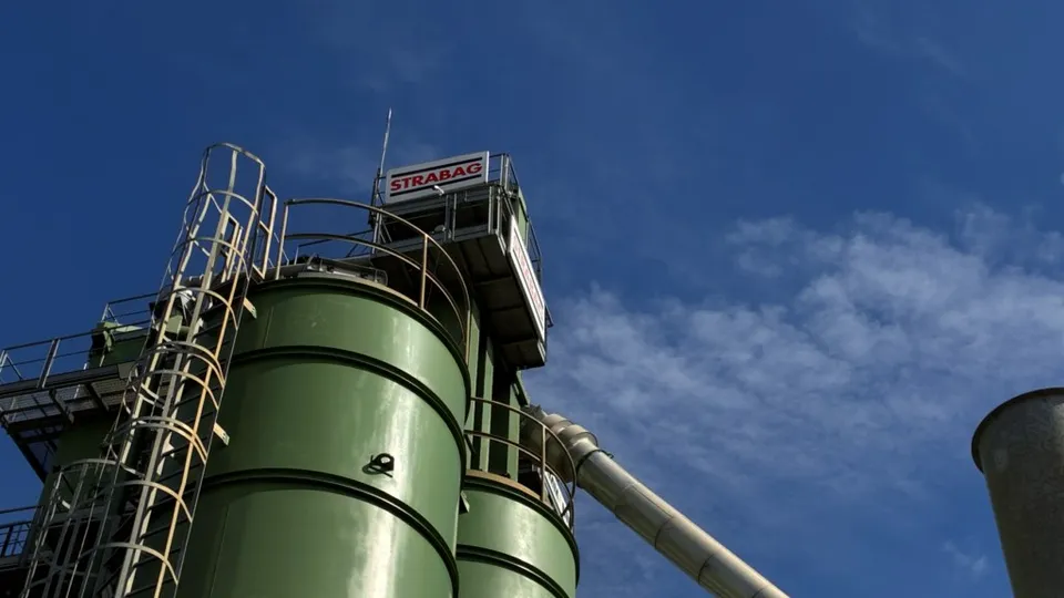 Două silozuri cilindrice verzi sunt situate sub un cer albastru cu nori subțiri, lângă o structură metalică înaltă cu un logo roșu și alb.
